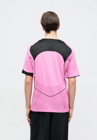 Μπλουζάκι αθλητικής εφαρμογής με κοντά μανίκια σε έντονο ροζ χρώμα, με μαύρες πλέγματος λεπτομέρειες, καμπυλωτές ραφές και αντίθετες ραφές στους ώμους και στα πλάγια.