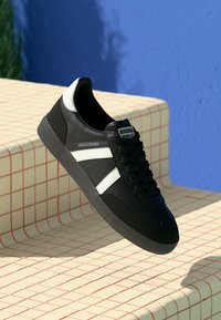 Black sneaker with white stripes and heel detail, "Jack & Jones" branding, displayed on cream tiled steps against a blue wall.