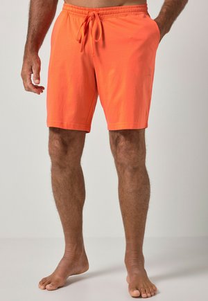 Mann trägt leuchtend orangefarbene Shorts mit Kordelzug, steht barfuß mit einer Hand in der Tasche vor einfarbigem Hintergrund.