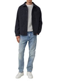 Marineblaue Wolljacke mit vorderen Knöpfen und Tasche, getragen über einem hellgrauen Strickpullover, kombiniert mit hellblauen Jeans und weißen Sneakers.