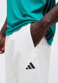 Άτομο που φοράει λευκό παντελόνι Adidas με τσέπη με φερμουάρ και πετρόλ μπλούζα με μαύρες ρίγες, χέρι στην τσέπη.