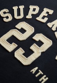Tejido negro con bordado en relieve de color crema. El texto incluye "SUPER" arriba de "23" y "ATH" en la parte inferior. Las texturas son suaves y detalladas.