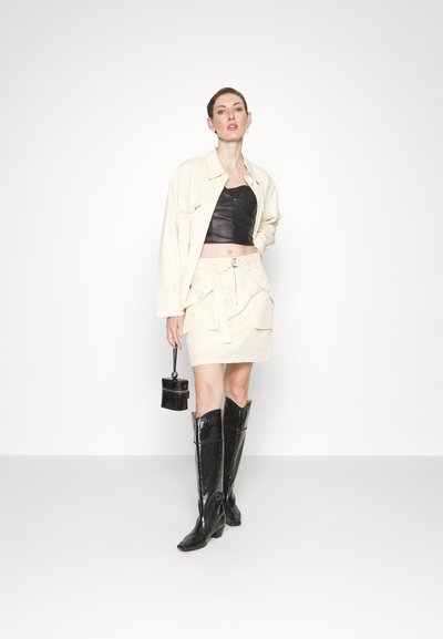 Veste oversize crème par-dessus un top court noir, associée à une jupe taille haute avec poches. Bottes noires arrivant au genou et un petit sac à main.