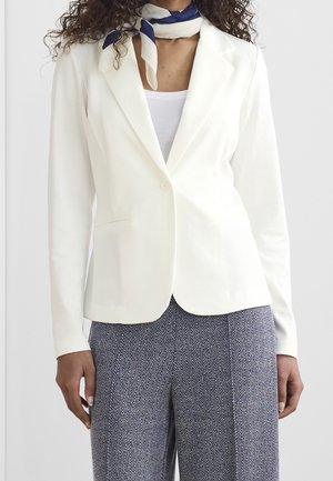 Frau trägt weißen Blazer über weißem Oberteil, gemusterte graue Hose und blau-weißes Halstuch um den Hals gebunden.