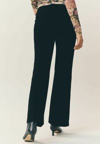 Kobieta ubrana w czarne szerokie spodnie z wysokim stanem, czarne botki na obcasie i kwiecistą bluzkę z długim rękawem, sfotografowana z tyłu.