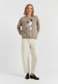 Pull beige avec un design graphique Snoopy, à manches longues et col rond. Associé à un pantalon large crème et des mocassins noirs.