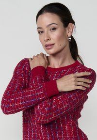 Pull en motif rouge avec un design tricoté texturé, comportant des accents bleus et crème ainsi que des poignets côtelés. Le modèle pose les bras croisés.