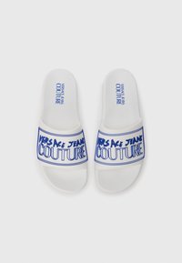 White slide sandals with a textured footbed, featuring a blue graphic print “VERSACE JEANS COUTURE” on the strap.