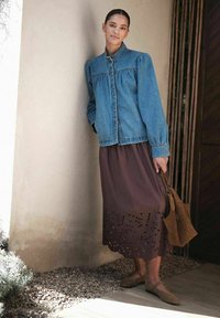 Kvinde lænende sig mod væggen iført en blå denimjakke, brun midi-nederdel med udskæringsdetaljer, holdende en brun ruskindtaske og flade sko.