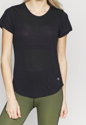 Frau trägt ein eng anliegendes schwarzes, strukturiertes Kurzarmshirt und grüne gestreifte Leggings, steht mit entspannten Armen seitlich am Körper.