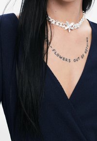 Top envolvente de color azul marino con textura acanalada, que presenta un collar choker de cadena plateada con un acento floral, y un tatuaje de texto visible en la zona de la clavícula.