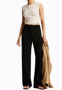 Top corto de encaje crema con mangas cap, combinado con pantalones de talle alto, de pierna ancha, negro. Capa exterior marrón sostenida en la mano. Sandalias planas.