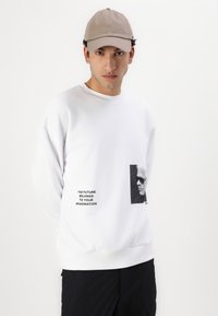 Hvit sweatshirt med rund hals og lange ermer, med en trykt svart grafikk og tekst på forsiden, kombinert med svarte shorts og en beige caps.