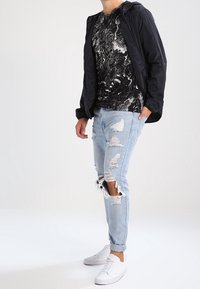 Svart mönstrad t-shirt, lätt navy jacka, slitna ljusblå jeans, vita sneakers. Avslappnad outfit med blandade texturer och färger.