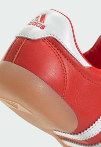 Crvena kožna cipela s bijelim jastučićem na peti i srebrnim detaljima. Ima glatku teksturu i poluprozirnu gumenu potplat.