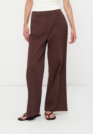 Femme portant un pantalon marron ample avec des poches latérales et une ouverture boutonnée sur le devant, associé à des sandales tongs marron foncé sur fond blanc.