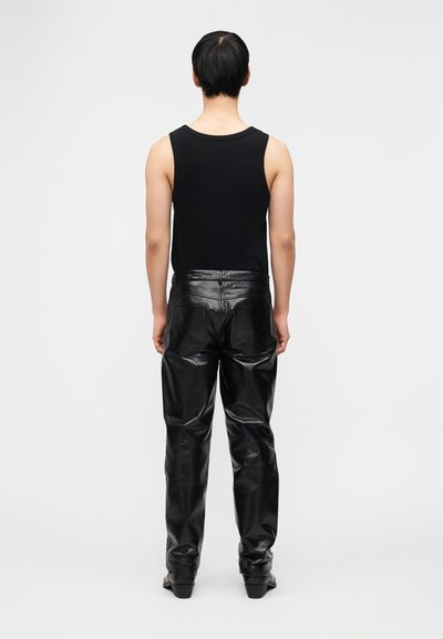 Άτομο που στέκεται με την πλάτη, φοράει μαύρο αμάν ικο τοπ και γυαλιστερό μαύρο δερμάτινο παντελόνι, σε απλό φόντο.