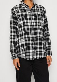 Camicia in flanella a quadri bianco e nero con maniche lunghe, chiusura a bottoni sul davanti e taschino sul petto, caratterizzata da una vestibilità morbida e una texture delicata.