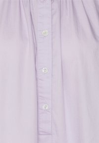 Camisa de botões lilás claro com uma textura suave de algodão, apresentando uma abertura vertical com botões brancos e um corte descontraído.