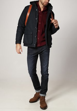 Hombre con chaqueta negra con capucha sobre suéter color vino, vaqueros oscuros con puños, botas marrones con cordones y correas de mochila de cuero marrón.
