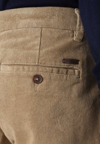 Beige corduroybroek met een achterzak voorzien van een bruine knoop en een klein metalen label met de tekst "ONLY & SONS." Gepoederde stof met geribbelde structuur.