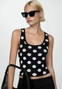 Top nero senza maniche con grandi pois bianchi; tessuto a maglia testurizzato. Indossato con occhiali da sole neri e una borsa nera.