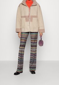 Flauschige beige Jacke mit strukturierter Oberfläche, kombiniert mit bunten gemusterten Schlaghosen. Hält eine kleine, runde lila Handtasche.