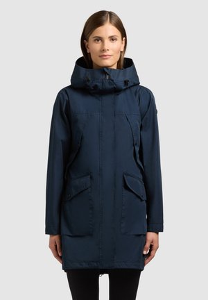 Žena oblečená v námornícky modrej kapucňovej parke so zipsom vpredu, klopovými vreckami a nastaviteľnými sťahovacími šnúrkami, stojaca proti jednoliatemu svetlému pozadiu.