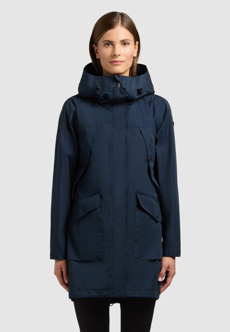 Femme portant une parka à capuche bleu marine avec fermeture éclair à l'avant, poches à rabat et cordons de serrage réglables, debout devant un fond clair uni.