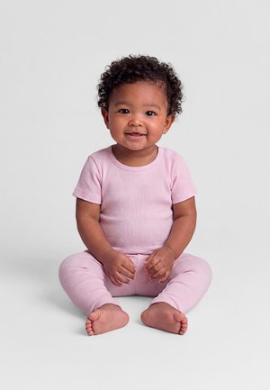 Copil mic zâmbitor cu păr creț, purtând un tricou roz cu mâneci scurte, cu dungi, și pantaloni asortați, șezând desculț pe o suprafață gri deschis.