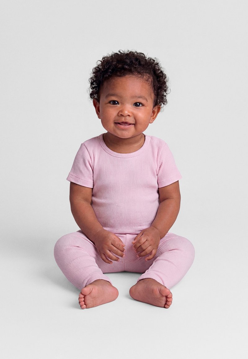 Bambino sorridente con capelli ricci, indossa una maglietta rosa a maniche corte a coste e pantaloni abbinati, seduto a piedi nudi su una superficie grigio chiaro.