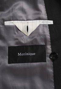 Étiquette en tissu noir avec logo blanc Matinique cousue sur la doublure grise du costume près d’un bouton sombre et détail d’une boucle en tissu rayé.