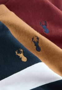 Trīs teksturēti džemperi tumši zaļā, zeltaini brūnā un tumši sarkanā krāsā, katrs ar izšūtu briedi emblēmu kontrastējošās krāsās.