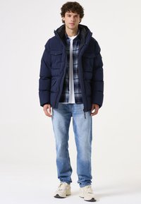Marineblauwe pufferjack met een hoge kraag, ritszakken en een gewatteerde textuur, gelaagd over een geruit overhemd en een witte t-shirt, gecombineerd met lichte jeans en sneakers.