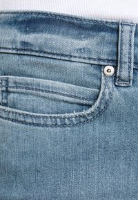 Jeans de denim azul claro con un diseño clásico de cinco bolsillos, que presentan costuras visibles, un cierre de botón metálico y una textura deslavada.
