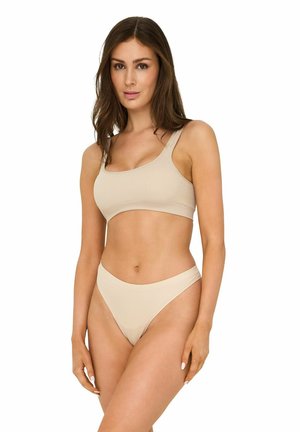 Woman with long brown hair wearing beige ribbed crop top and matching high-cut bikini bottoms standing against white background.