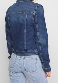 Tmavě modrá džínová bunda s přiléhavým střihem, kontrastním prošíváním a knoflíkovými detaily na rukávech; zobrazená zezadu.