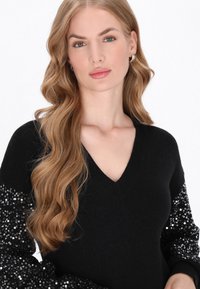 Zwarte trui met V-hals, met textuur op de zwarte pailletten mouwen. Zacht materiaal, ontworpen voor een aansluitende pasvorm en een gladde afwerking.