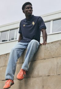 Marineblaues Poloshirt mit goldenem Logo und geometrischem Muster, hellblaue Jeans, orangefarbene Sneaker mit grünen Streifen. Modell sitzt auf einer Betonschicht.