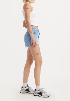 Femme debout de profil portant un crop top blanc, un short en denim bleu clair, des chaussettes blanches, des baskets argentées et plusieurs petits tatouages sur les bras et les jambes.