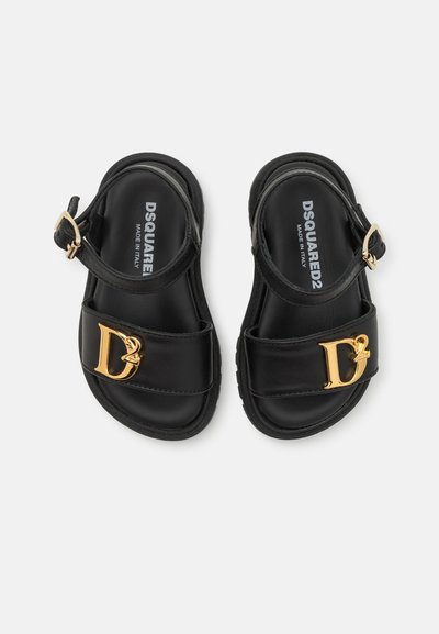 Sandales en cuir noir avec deux larges sangles, accents de logo doré, boucle réglable à la cheville et semelle texturée. Fabriquées en Italie.
