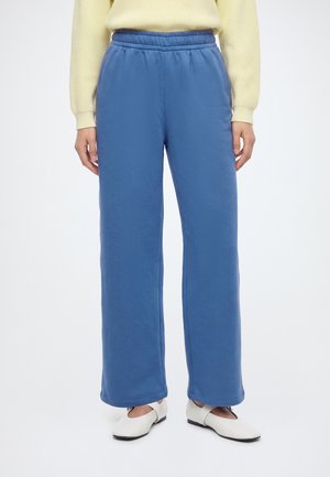 Persona con pantalones azules de pierna ancha, un suéter amarillo claro y zapatos planos blancos con tiras, de pie frente a un fondo liso.