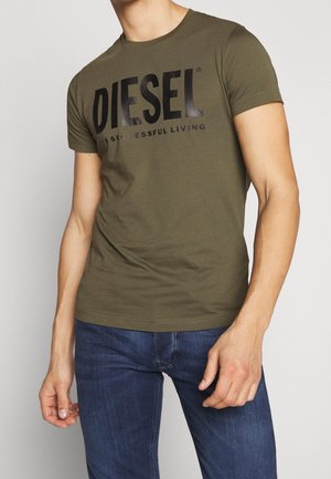 Mann trägt ein olivgrünes Diesel-T-Shirt mit schwarzem Logotext und blaue Jeans, steht vor einem schlichten weißen Hintergrund.