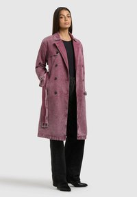 Donna in piedi che indossa un lungo trench doppiopetto viola sbiadito, top nero, pantaloni neri a gamba larga e scarpe nere con punta, su sfondo grigio.