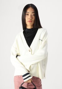 Cardigan bianco lavorato a maglia con scollatura a V profonda, orlo e polsini a coste con strisce blu navy. Chiusura con un bottone e dettaglio del logo sono visibili.