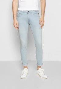 Jeans ajustados de color azul claro con una textura ligeramente desgastada, que cuentan con un cierre de botón y puños enrollados, combinados con zapatillas blancas.