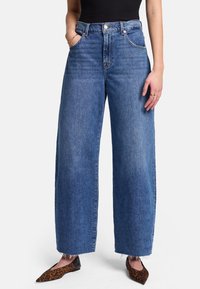 Jeans de pierna ancha en denim azul medio, de cintura alta con bolsillos frontales, dobladillo deshilachado y un ligero desvanecimiento. Combinados con bailarinas de punta afilada con estampado de leopardo.