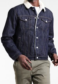 Dark denim jacket with a shearling collar; features two chest pockets, button closures, and contrasting stitching. Worn over light-colored pants.