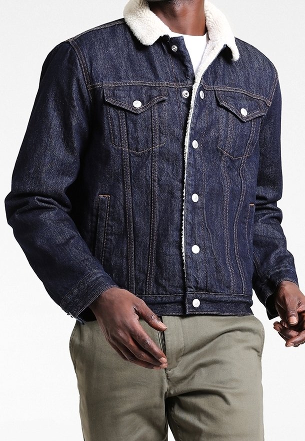 Dark denim jacket with a shearling collar; features two chest pockets, button closures, and contrasting stitching. Worn over light-colored pants.