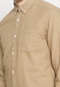 Braunes Hemd mit Knöpfen aus strukturiertem Stoff, ausgestattet mit einem klassischen Kragen, weißen Knöpfen und einer einzelnen Brusttasche.